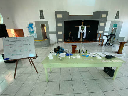 Photo of a table set up for a community science workshop on DNA sequencing. To the left of the table is a whiteboard with a diagram explaining PCR. The setting is a church.