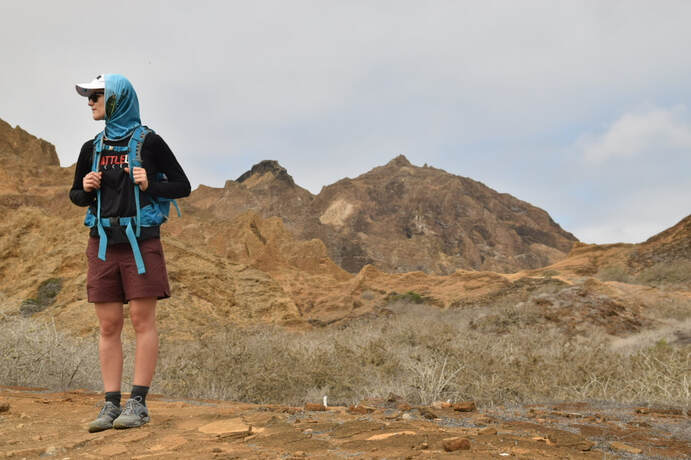 Photo of Jacqui in hiking attire standing to the left of the frame, on dirt with brush and with rocky, yellow cliffs behind. The sky is cloudy with only a hint of blue.